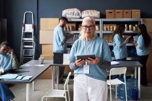 Portrait of senior woman holding tablet in office with colleagues working in background. Shelves filled with supplies and modern office furniture creating professional atmosphere
